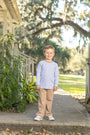 A young boy stands on a concrete walkway outdoors, wearing a light lavender long-sleeve shirt and tan pants.