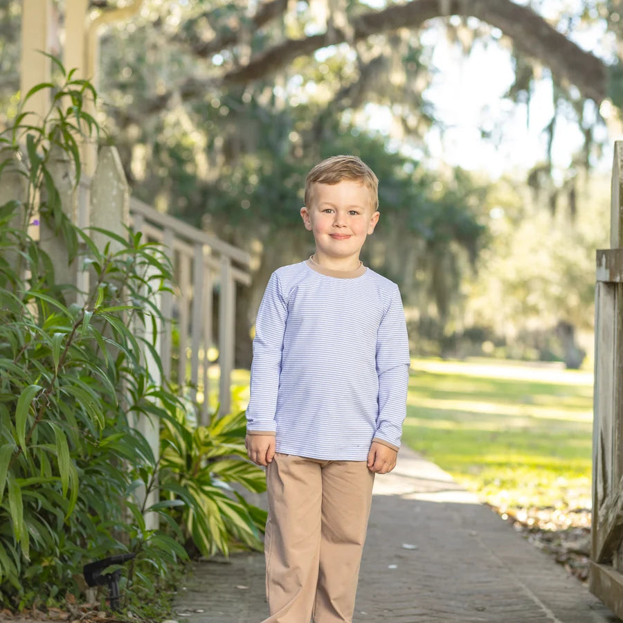 A young boy stands on a concrete walkway outdoors, wearing a light lavender long-sleeve shirt and tan pants.
