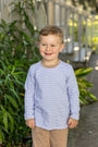 A young boy smiling outdoors, wearing a light blue and white striped long-sleeve shirt with tan pants.