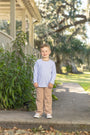 A young boy stands on a stone walkway in front of a house porch, wearing a light periwinkle blue long-sleeve crewneck shirt and tan pants.