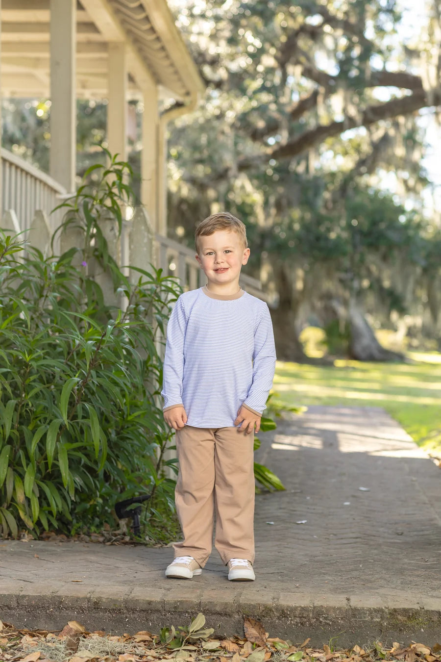 A young boy stands on a stone walkway in front of a house porch, wearing a light periwinkle blue long-sleeve crewneck shirt and tan pants.