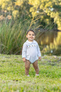 A baby standing on grass outdoors near tall reeds and water, wearing a light blue long-sleeve romper with small embroidered details across the chest.