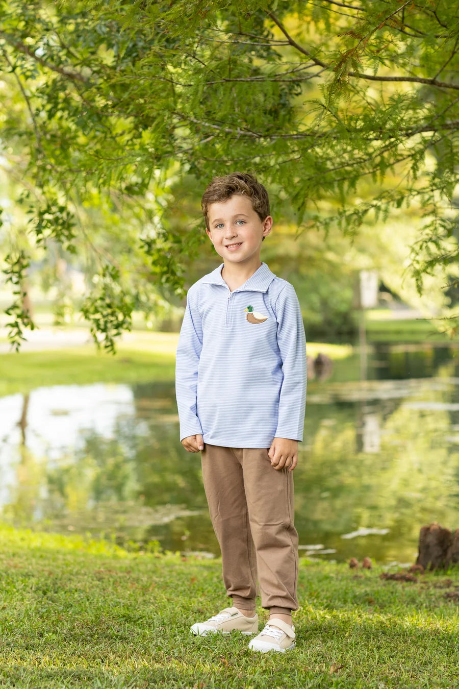 A young boy stands on a grassy lawn in front of a pond, wearing a light blue long-sleeve polo shirt with a small duck logo on the left chest, brown pants, and white sneakers.