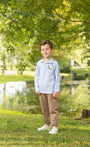 A young boy stands on a grassy lawn in front of a pond, wearing a light blue long-sleeve polo shirt with a small duck logo on the left chest, brown pants, and white sneakers.