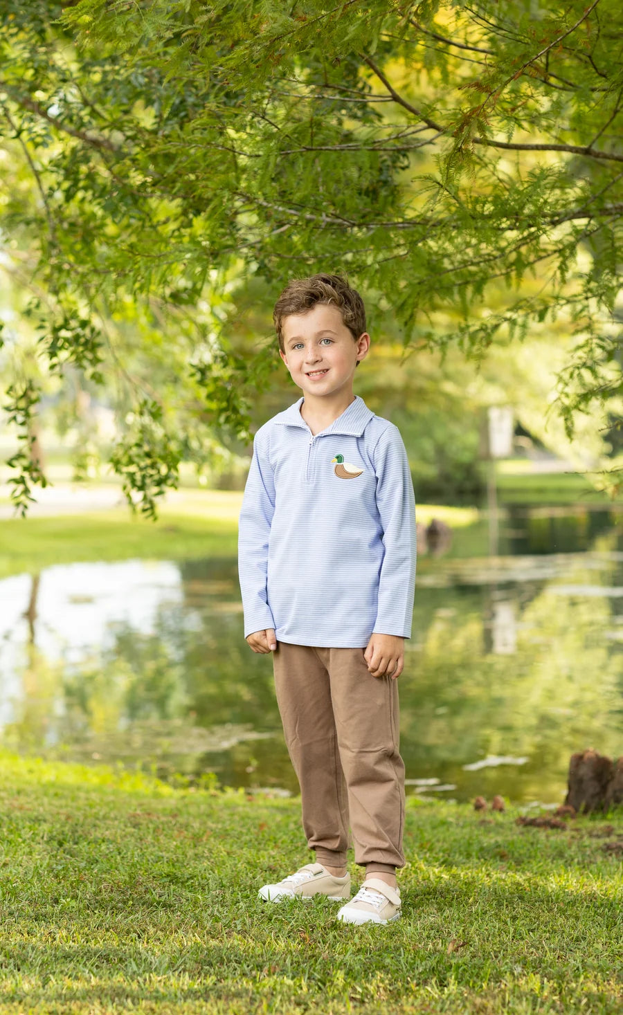 A young boy stands on a grassy lawn in front of a pond, wearing a light blue long-sleeve polo shirt with a small duck logo on the left chest, brown pants, and white sneakers.