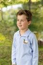 A young boy wearing a light blue and white striped long-sleeve polo shirt with a green and yellow duck patch on the chest, standing outdoors in a grassy, wooded area.