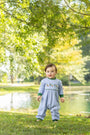 A baby standing outdoors on grass near a pond under leafy trees, wearing a light blue long-sleeve footless romper with green trim and small embroidered details on the chest.