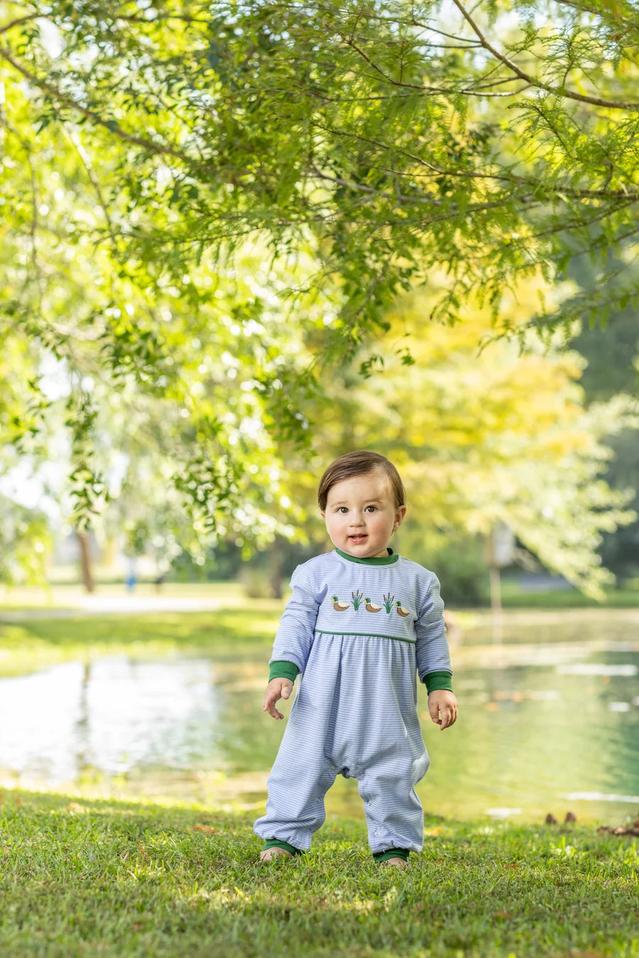 A baby standing outdoors on grass near a pond under leafy trees, wearing a light blue long-sleeve footless romper with green trim and small embroidered details on the chest.