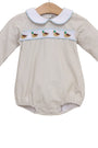 A baby long-sleeve romper in a light beige fabric, featuring a white Peter Pan collar with light blue trim, a horizontal band of embroidered ducks across the chest, and light blue piping along the cuffs and collar, hanging on a wooden hanger against a white background.