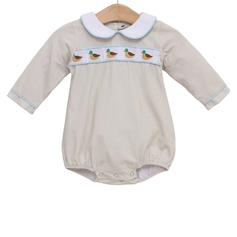 A baby long-sleeve romper in a light beige fabric, featuring a white Peter Pan collar with light blue trim, a horizontal band of embroidered ducks across the chest, and light blue piping along the cuffs and collar, hanging on a wooden hanger against a white background.