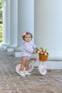 A toddler girl wearing a light lavender patterned romper and white sneakers sits on a pink tricycle with a wicker basket filled with tulips, positioned on a brick walkway in front of white classical columns.