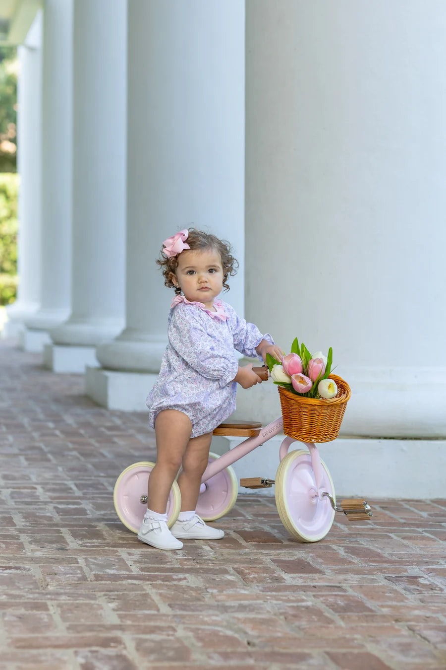 A toddler girl wearing a light lavender patterned romper and white sneakers sits on a pink tricycle with a wicker basket filled with tulips, positioned on a brick walkway in front of white classical columns.