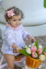 A toddler girl with curly brown hair wearing a light purple floral romper and a pink bow sits on a pink tricycle with a wicker basket filled with pink and white tulips.