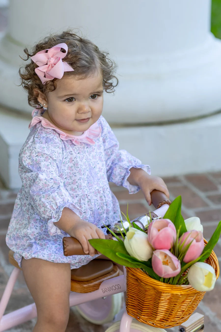 A toddler girl with curly brown hair wearing a light purple floral romper and a pink bow sits on a pink tricycle with a wicker basket filled with pink and white tulips.