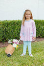 A young girl with blonde hair stands on a green lawn in front of a hedge, wearing a light pink long-sleeve dress with light blue trim and pockets, paired with light blue leggings. She is holding a small wicker basket filled with flowers.