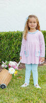 A young girl with blonde hair stands on a green lawn in front of a hedge, wearing a light pink long-sleeve dress with light blue trim and pockets, paired with light blue leggings. She is holding a small wicker basket filled with flowers.