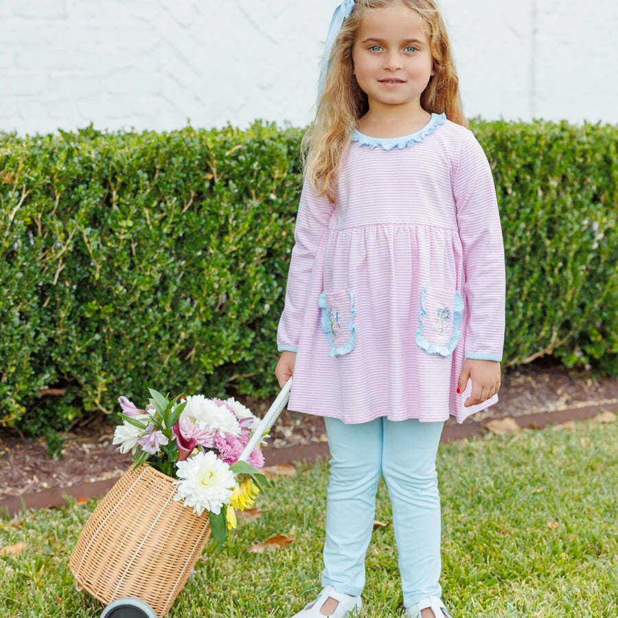 A young girl with blonde hair stands on a green lawn in front of a hedge, wearing a light pink long-sleeve dress with light blue trim and pockets, paired with light blue leggings. She is holding a small wicker basket filled with flowers.