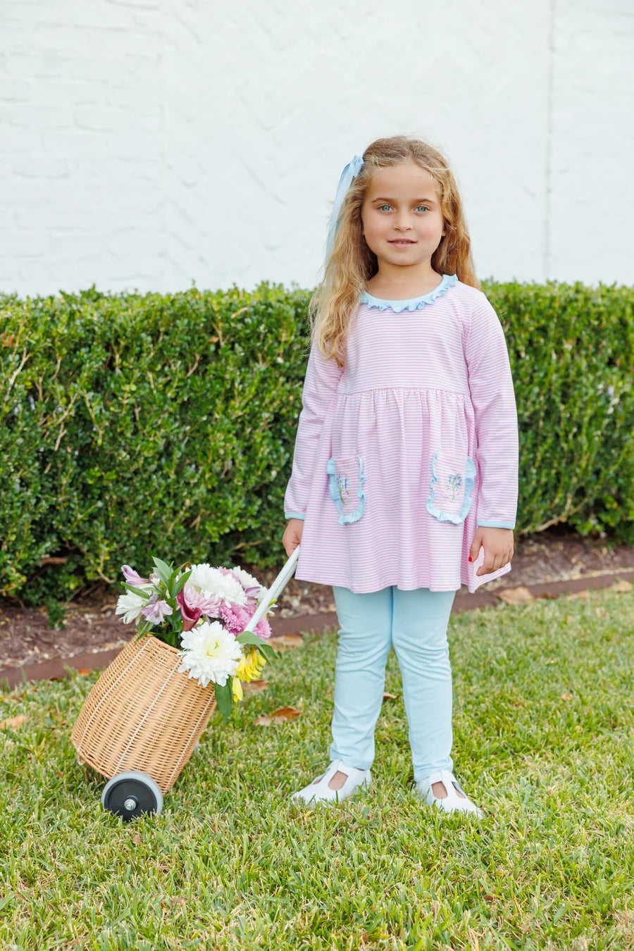 A young girl with blonde hair stands on a green lawn in front of a hedge, wearing a light pink long-sleeve dress with light blue trim and pockets, paired with light blue leggings. She is holding a small wicker basket filled with flowers.