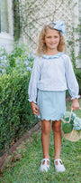 A young girl stands on a grassy lawn in front of a house, wearing a light blue and white vertically striped long-sleeve blouse with a ruffled collar, a light blue skirt, and white Mary Jane shoes. She is holding a small woven basket with flowers and wearing a matching light blue bow in her hair.