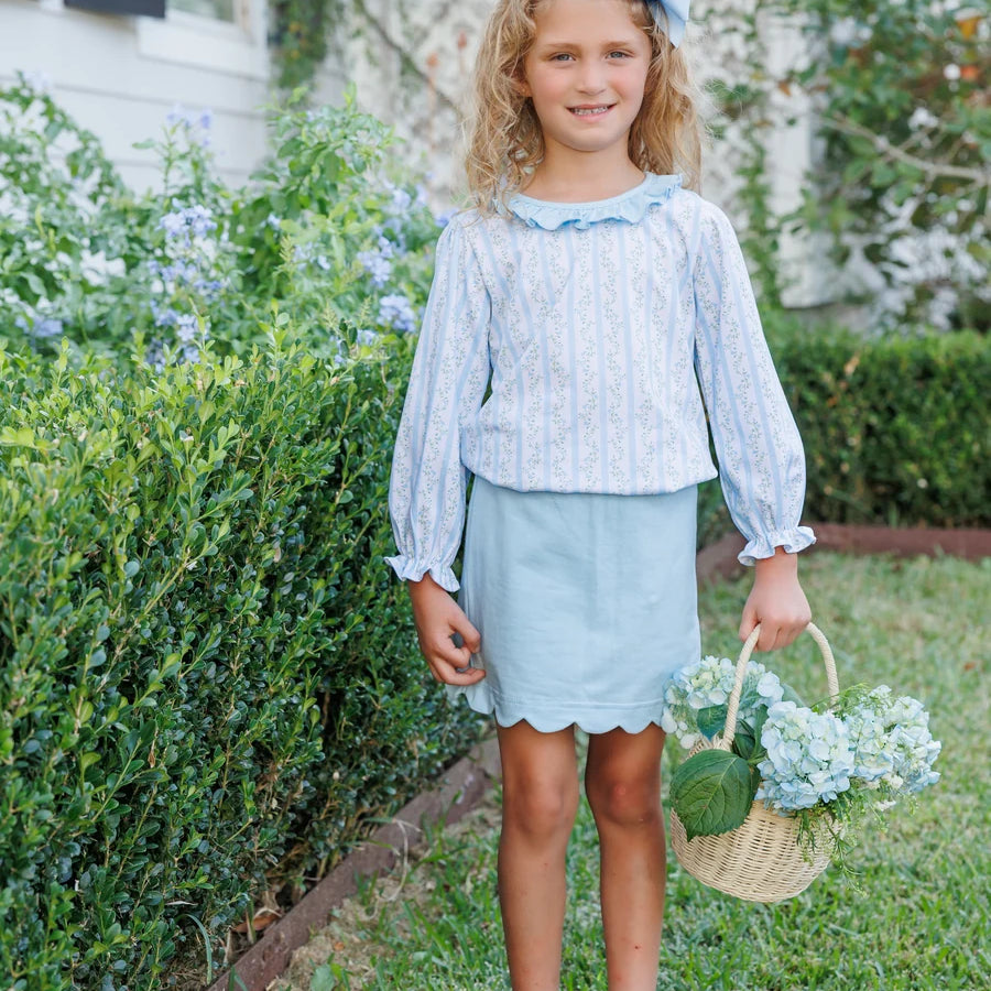 A young girl stands on a grassy lawn in front of a house, wearing a light blue and white vertically striped long-sleeve blouse with a ruffled collar, a light blue skirt, and white Mary Jane shoes. She is holding a small woven basket with flowers and wearing a matching light blue bow in her hair.