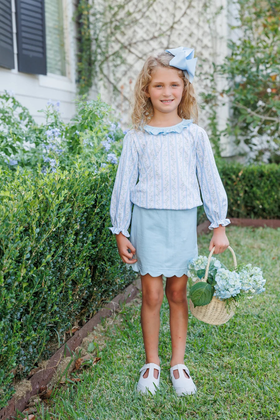 A young girl stands on a grassy lawn in front of a house, wearing a light blue and white vertically striped long-sleeve blouse with a ruffled collar, a light blue skirt, and white Mary Jane shoes. She is holding a small woven basket with flowers and wearing a matching light blue bow in her hair.