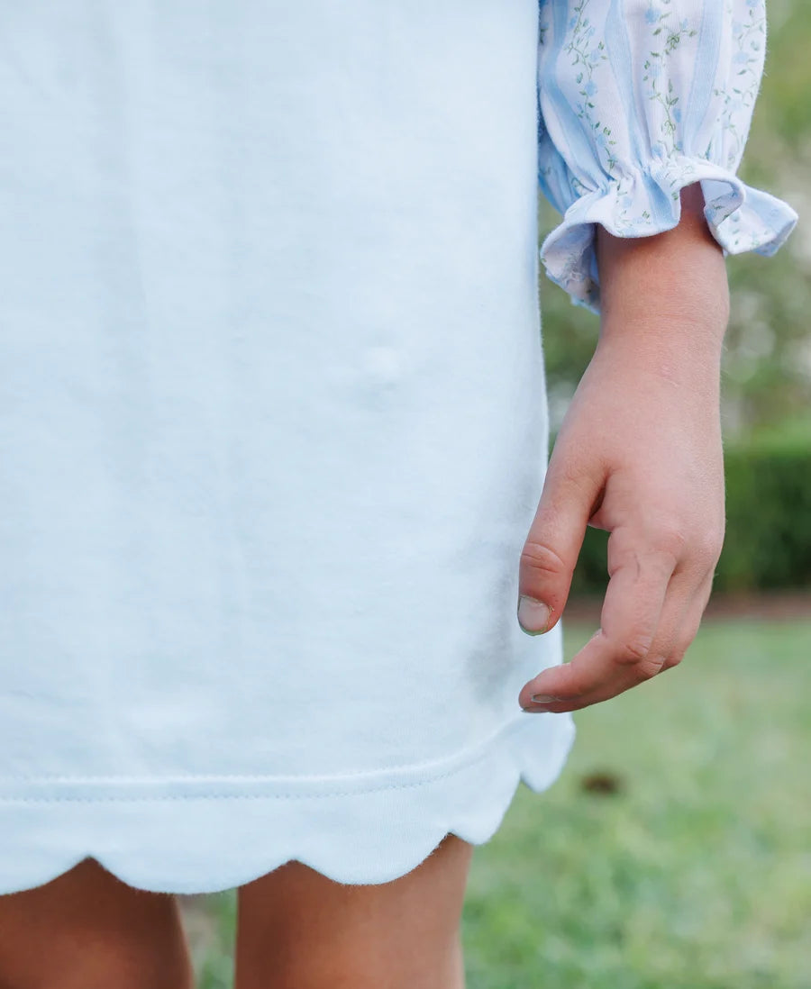 Close-up of a child wearing a light blue dress with a scalloped hem and a floral long-sleeve top, standing outdoors on grass.