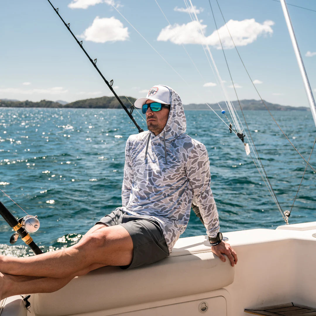 Man on a sailboat wearing sunglasses and a patterned shirt with a hood, sitting on the edge of the boat.