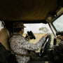 Person in camouflage attire driving a vehicle with binoculars, camera, and rifle, in a natural setting.
