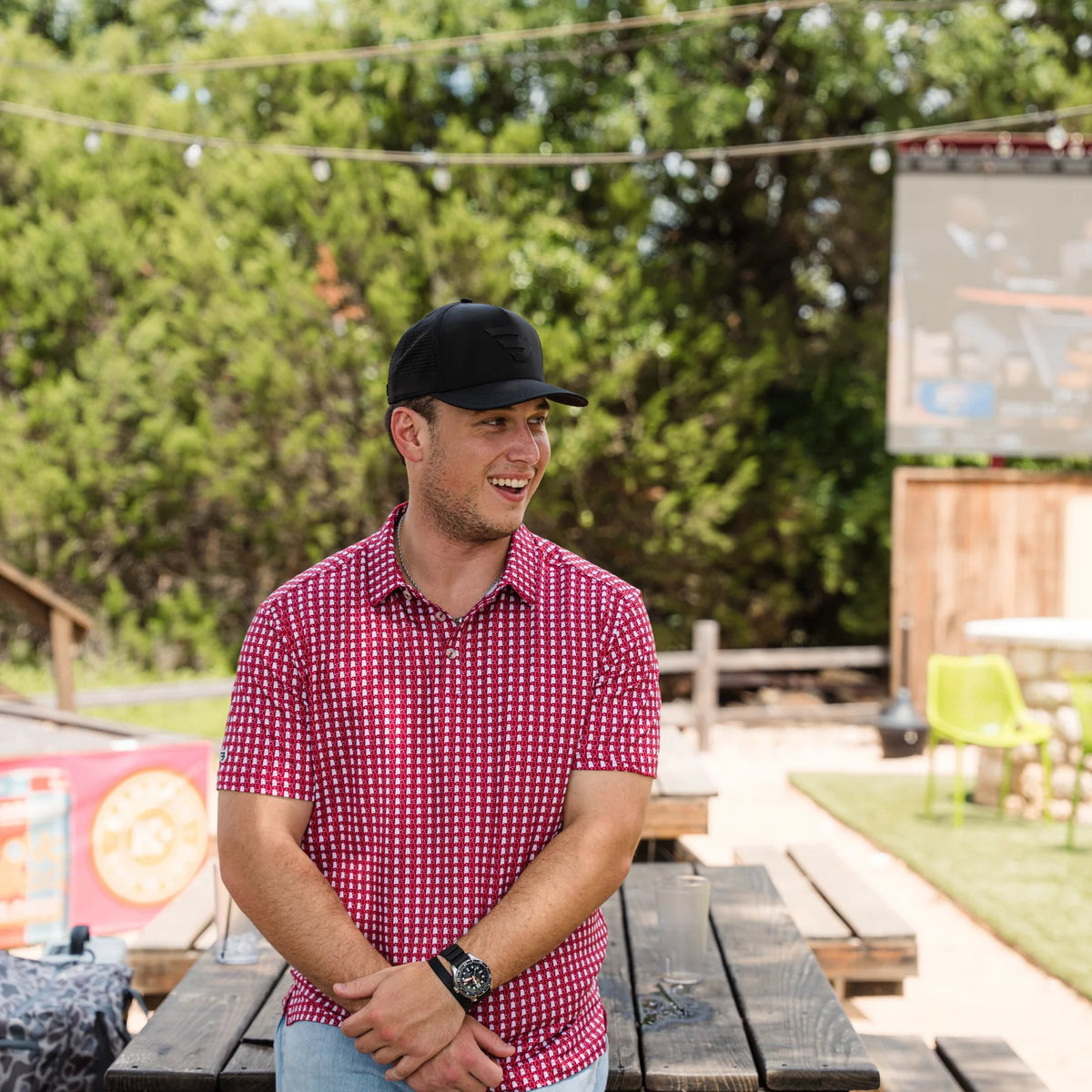 Man wearing a red checkered shirt and black cap standing outdoors with trees and a wooden structure in the background.