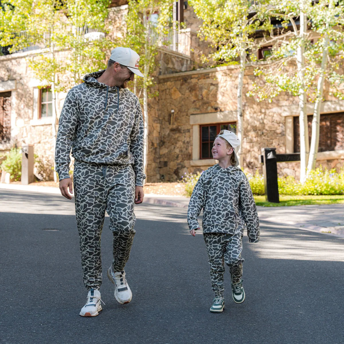 Man and child walking outdoors in matching camouflage outfits with a building in the background.
