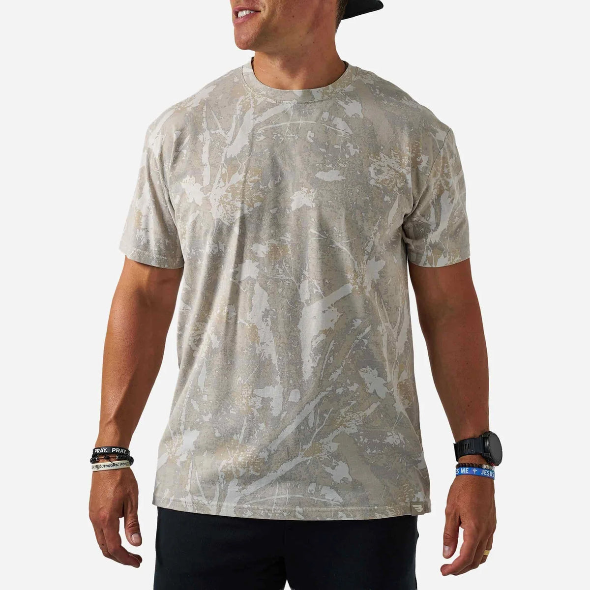 A man wearing a short-sleeve crewneck t-shirt with an abstract camouflage pattern in beige, gray, and white, paired with black pants and accessories.