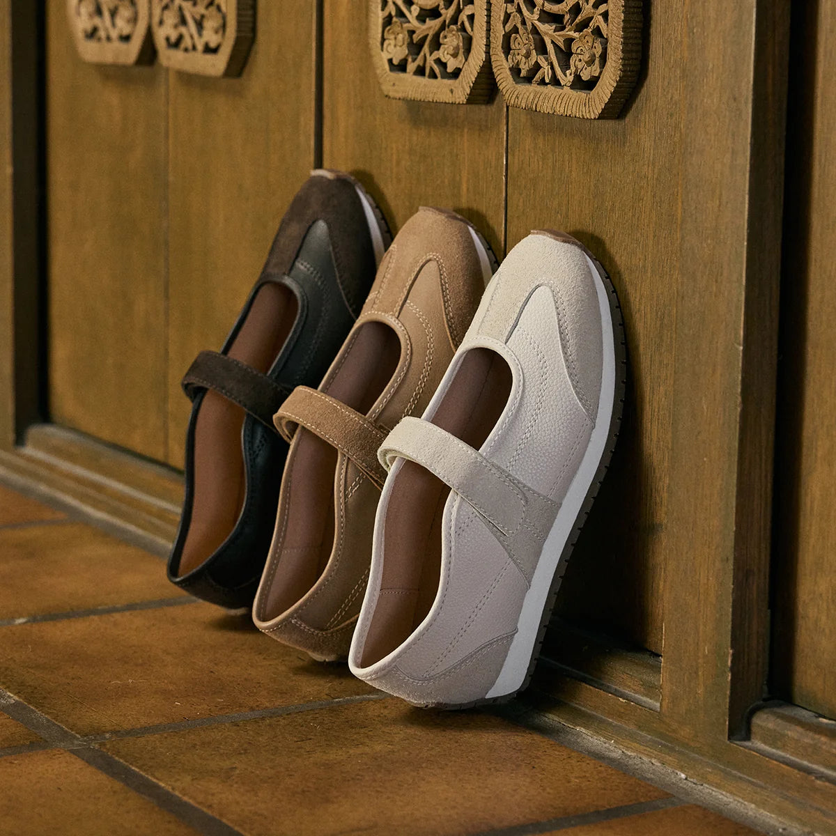 Three slip-on wedge shoes in black, tan, and light beige are lined up against a carved wooden door on a tiled floor.