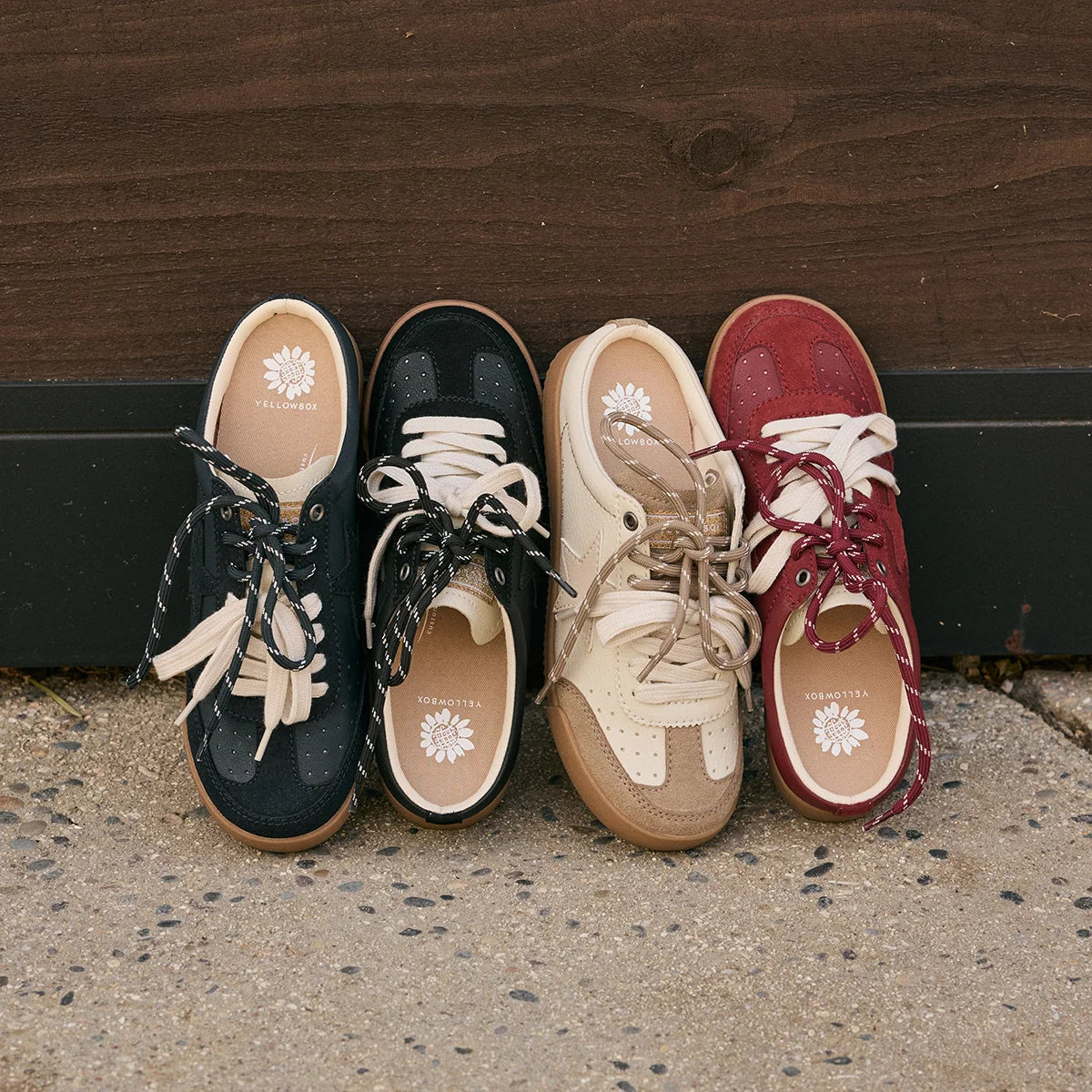 Four pairs of low-top lace-up sneakers arranged side by side on concrete against a dark wooden wall. The shoes are shown in four colorways: navy with cream, black with cream, beige with tan, and burgundy with cream.