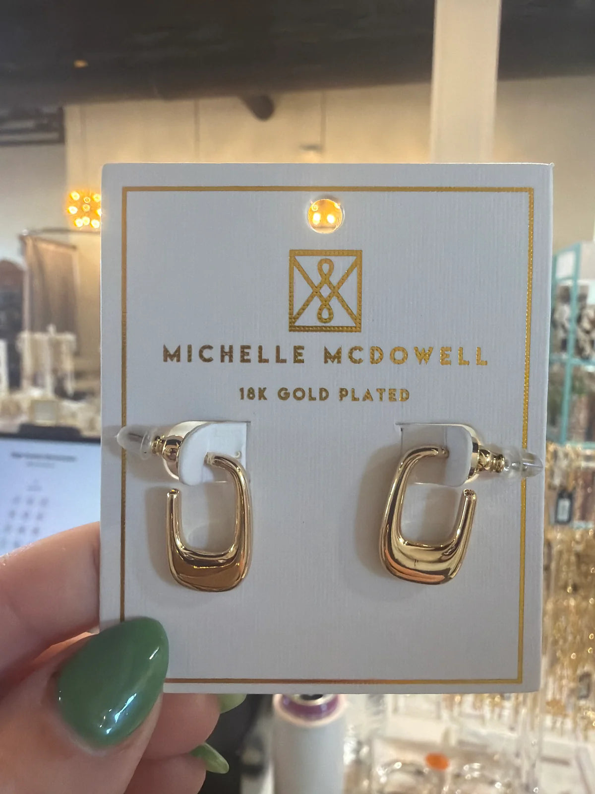 Gold hoop earrings on a Michelle McDowell branded card held by a hand with green nail polish.