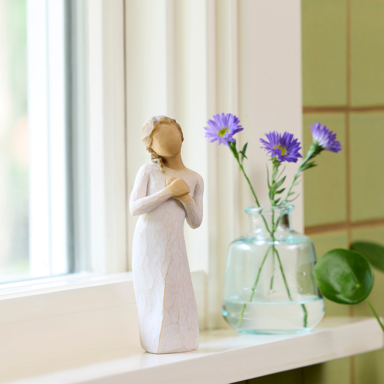 Statue of a woman holding flowers next to a vase with purple flowers on a windowsill.