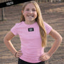 Young girl wearing a pink t-shirt with 'LULU MAC' logo outdoors.