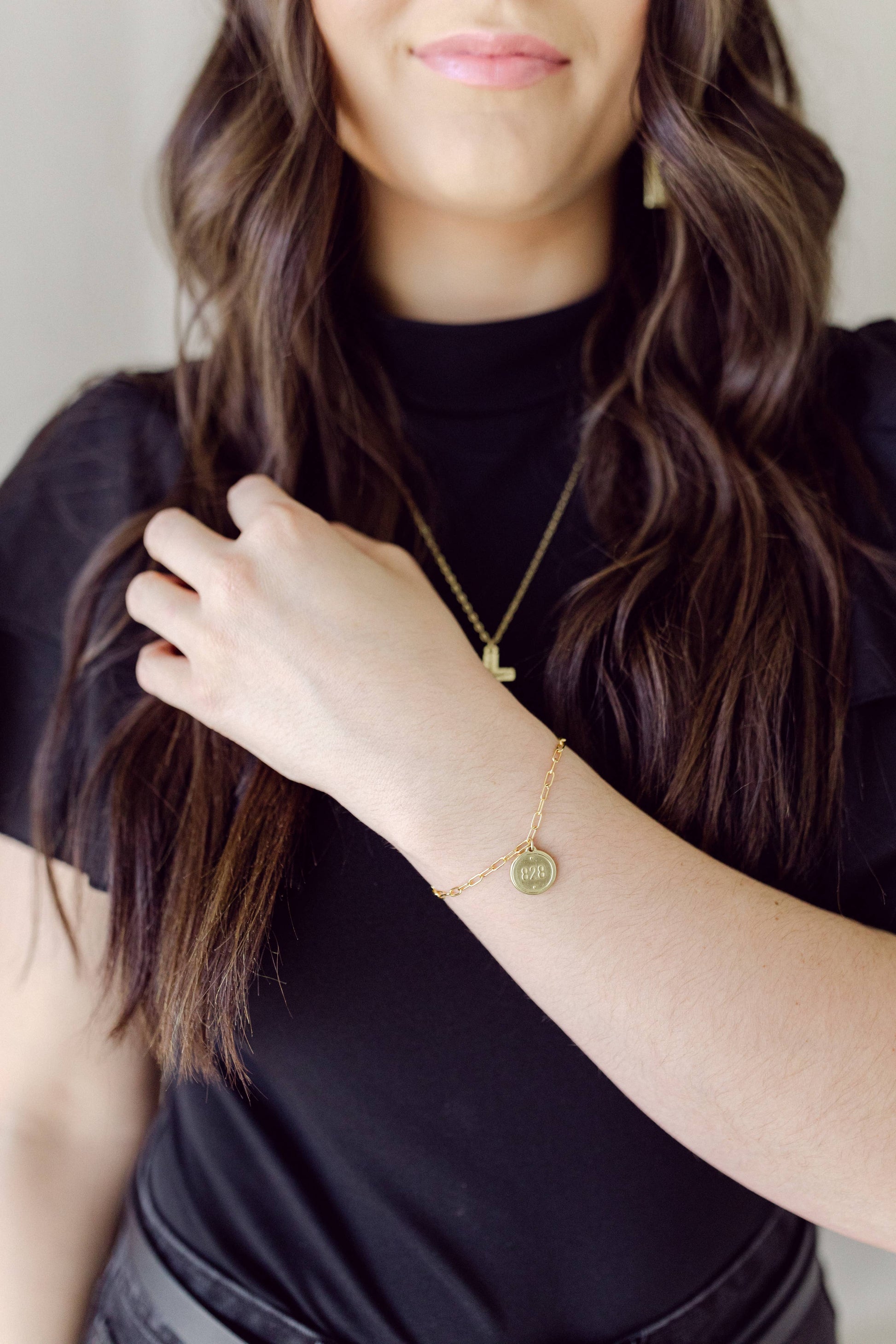 A close-up lifestyle shot of a woman wearing a delicate gold chain necklace and a matching gold bracelet with small circular charms, paired with a black t-shirt.