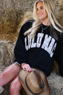 Woman sitting on hay wearing a black sweatshirt with 'CULU MAC' text, holding a cowboy hat.