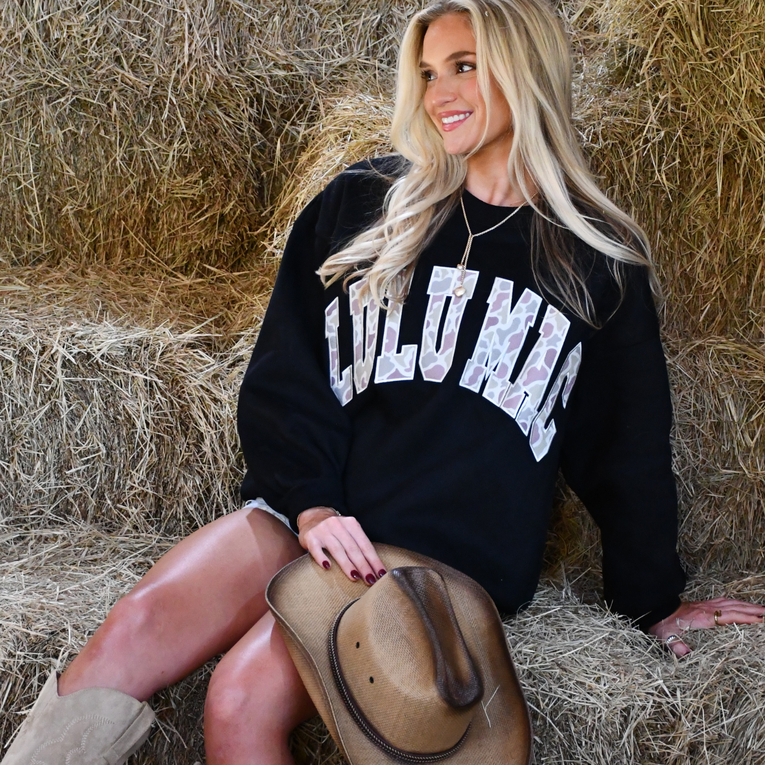 Woman sitting on hay wearing a black sweatshirt with 'CULU MAC' text, holding a cowboy hat.