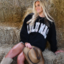 Woman sitting on hay wearing a black sweatshirt with 'CULU MAC' text, holding a cowboy hat.