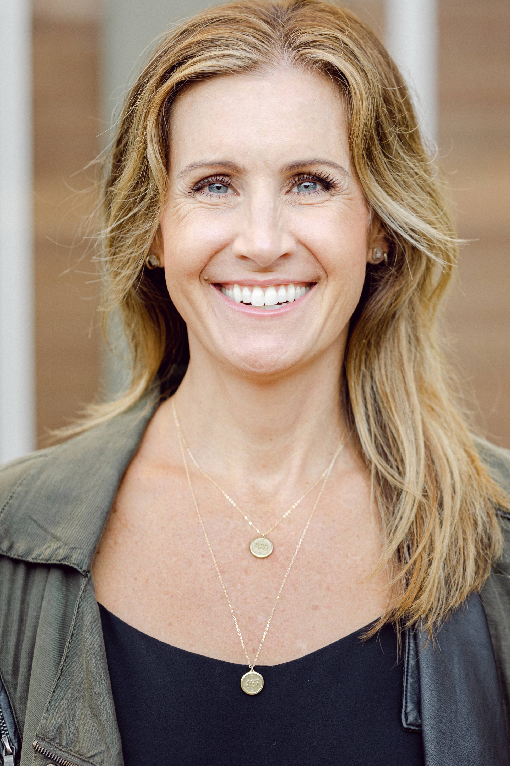 Woman with blonde hair wearing a green jacket and black top, smiling outdoors.