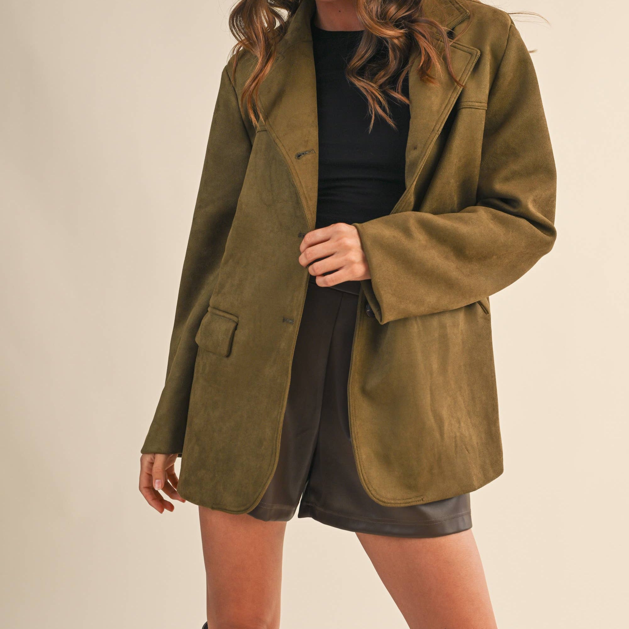 Woman wearing a green suede jacket over a black top and shorts on a plain background