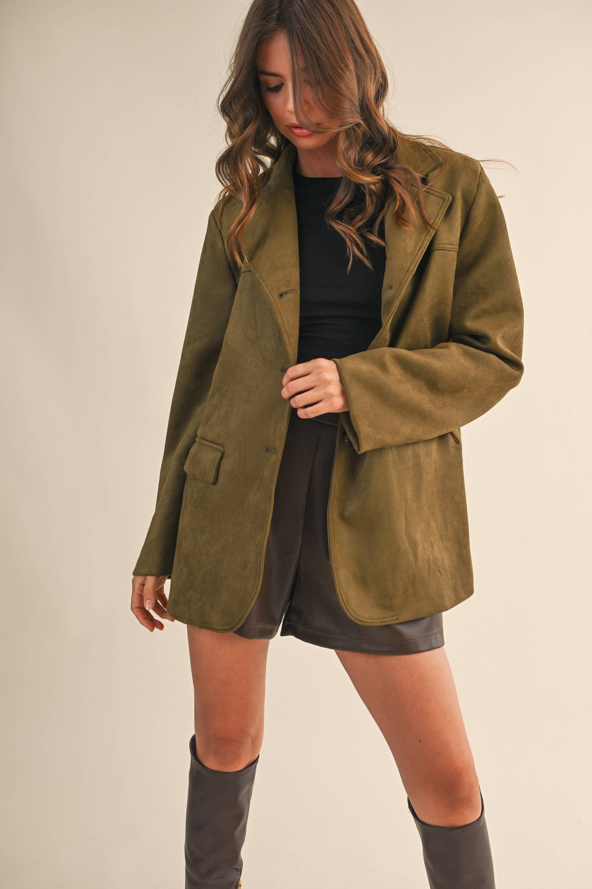 Woman wearing a green suede jacket over a black top and shorts on a plain background