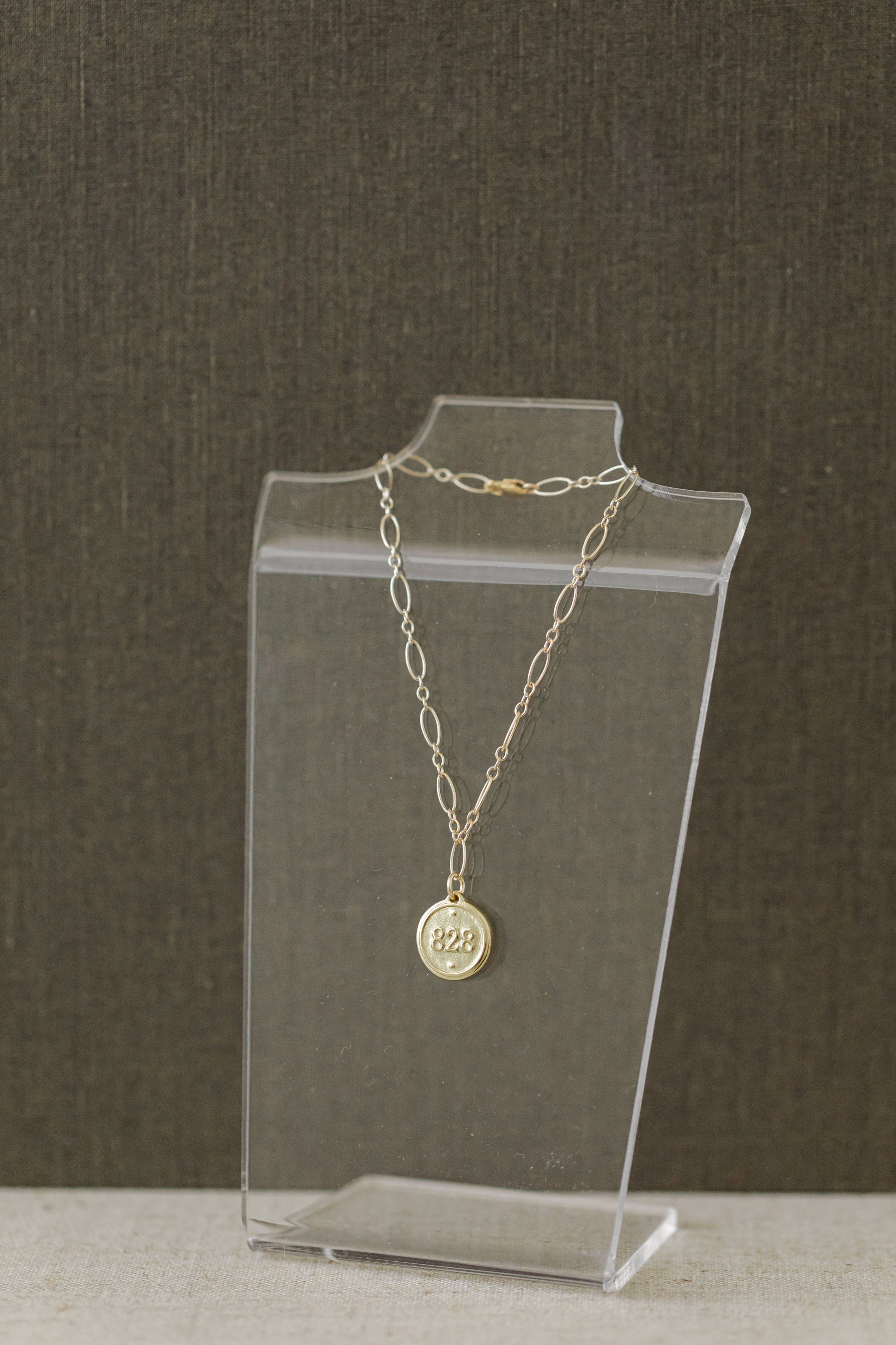 A delicate gold chain necklace with a round medallion pendant displaying '825' sterling silver purity stamp, displayed on a clear acrylic stand against a dark textured background.