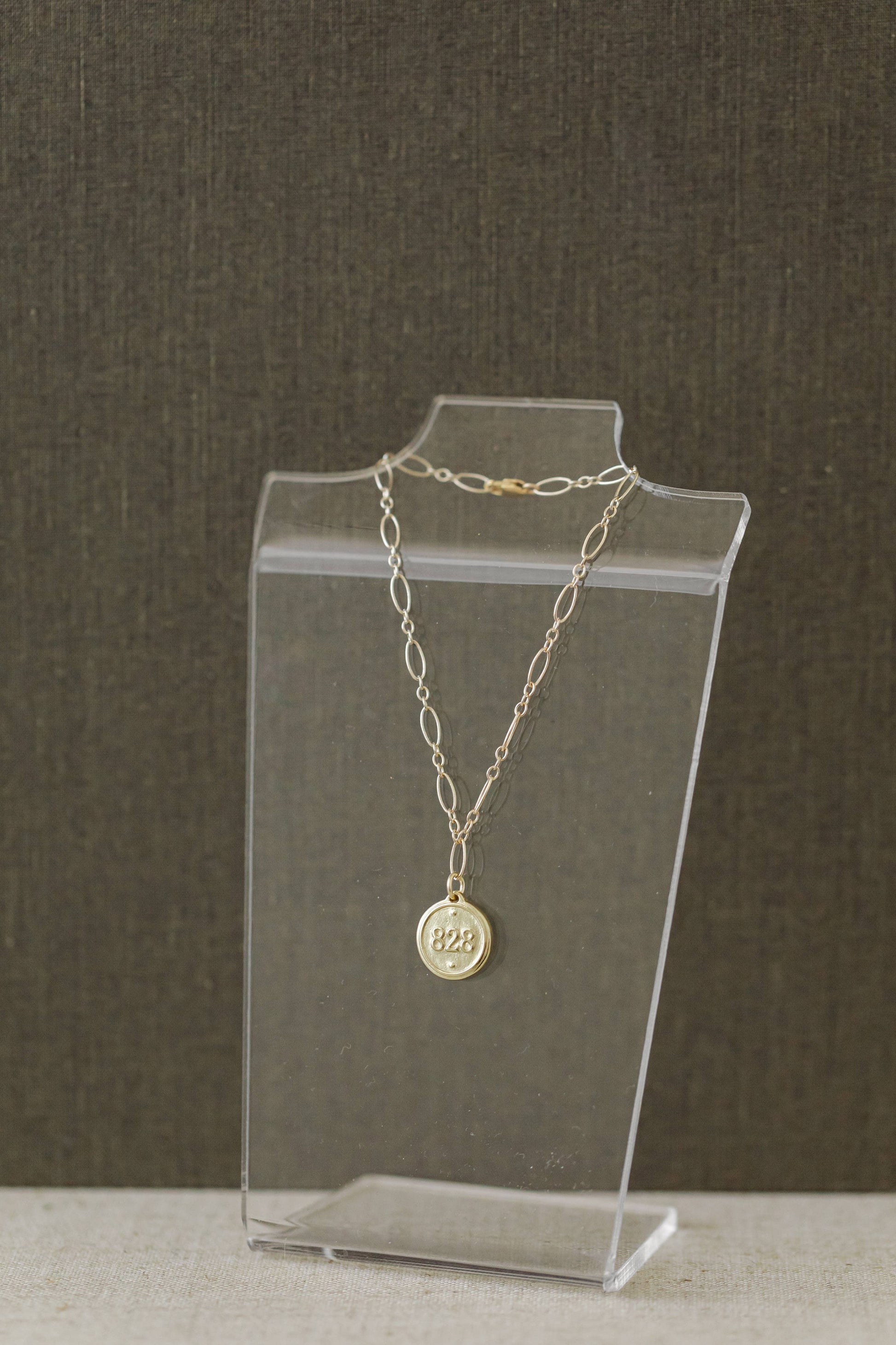 A delicate gold chain necklace with a round medallion pendant displaying '825' sterling silver purity stamp, displayed on a clear acrylic stand against a dark textured background.