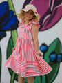 Young girl in a pink and white striped dress and sun hat standing in front of a colorful floral background.