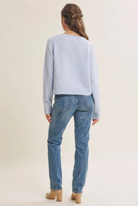 A woman shown from the back wearing a light blue knit sweater and blue jeans, standing against a plain light beige studio background.