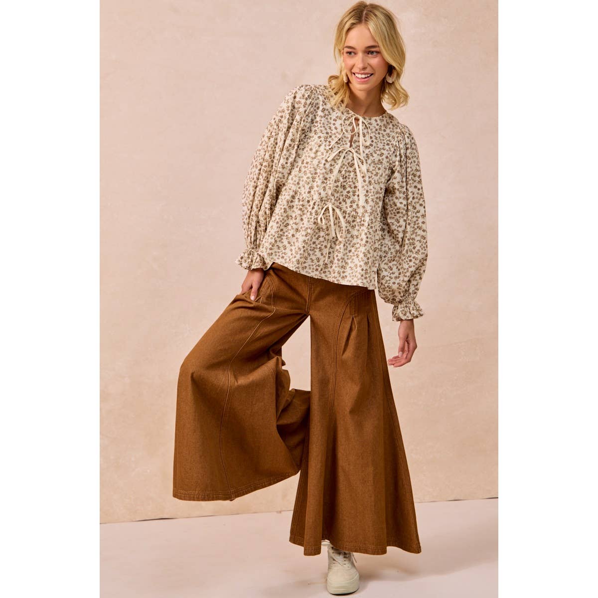 A woman modeling a beige and brown floral patterned long-sleeve blouse with a drawstring neckline, paired with wide-leg brown pants and white sneakers, set against a solid beige background.