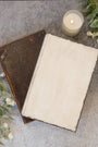 An open vintage-style book with a blank cream page lies on a textured gray surface, partially covering a closed dark brown book. A lit candle and white flowers are placed nearby as styled props.