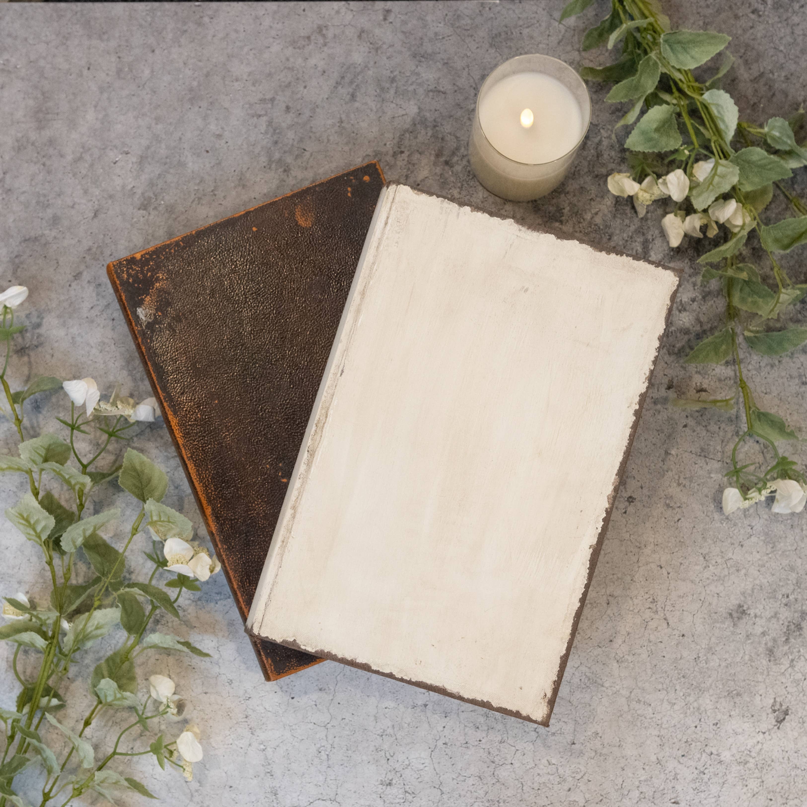 An open vintage-style book with a blank cream page lies on a textured gray surface, partially covering a closed dark brown book. A lit candle and white flowers are placed nearby as styled props.